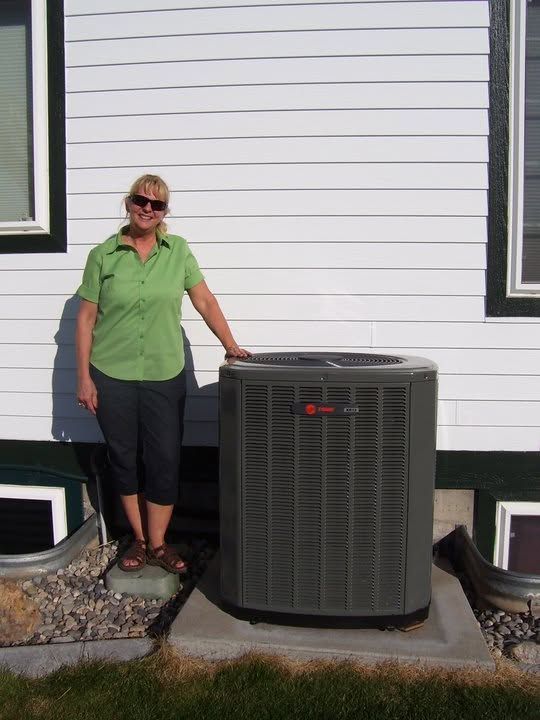 Woman standing beside outdoor HVAC unit against a white house wall.