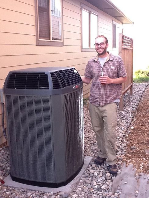 Man standing beside an outdoor HVAC unit next to a beige house, holding a drink on a gravel path.