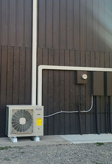 Outdoor air conditioning unit beside a dark building wall with white conduit piping.