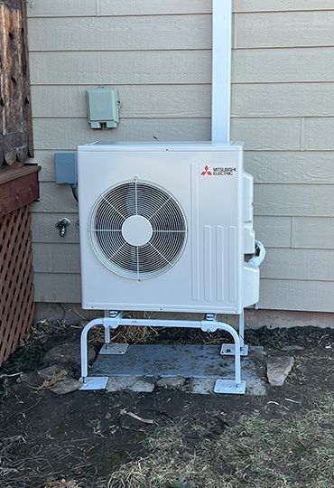 White Mitsubishi outdoor AC unit on a wall-mounted bracket beside a house wall.