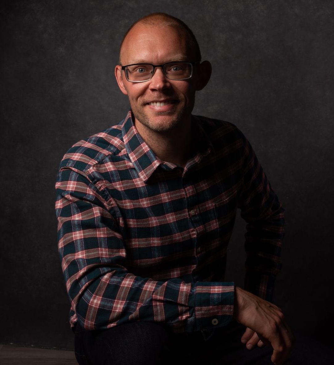 A man wearing glasses and a plaid shirt is sitting down and smiling.