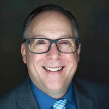 A man wearing glasses and a suit and tie is smiling for the camera.