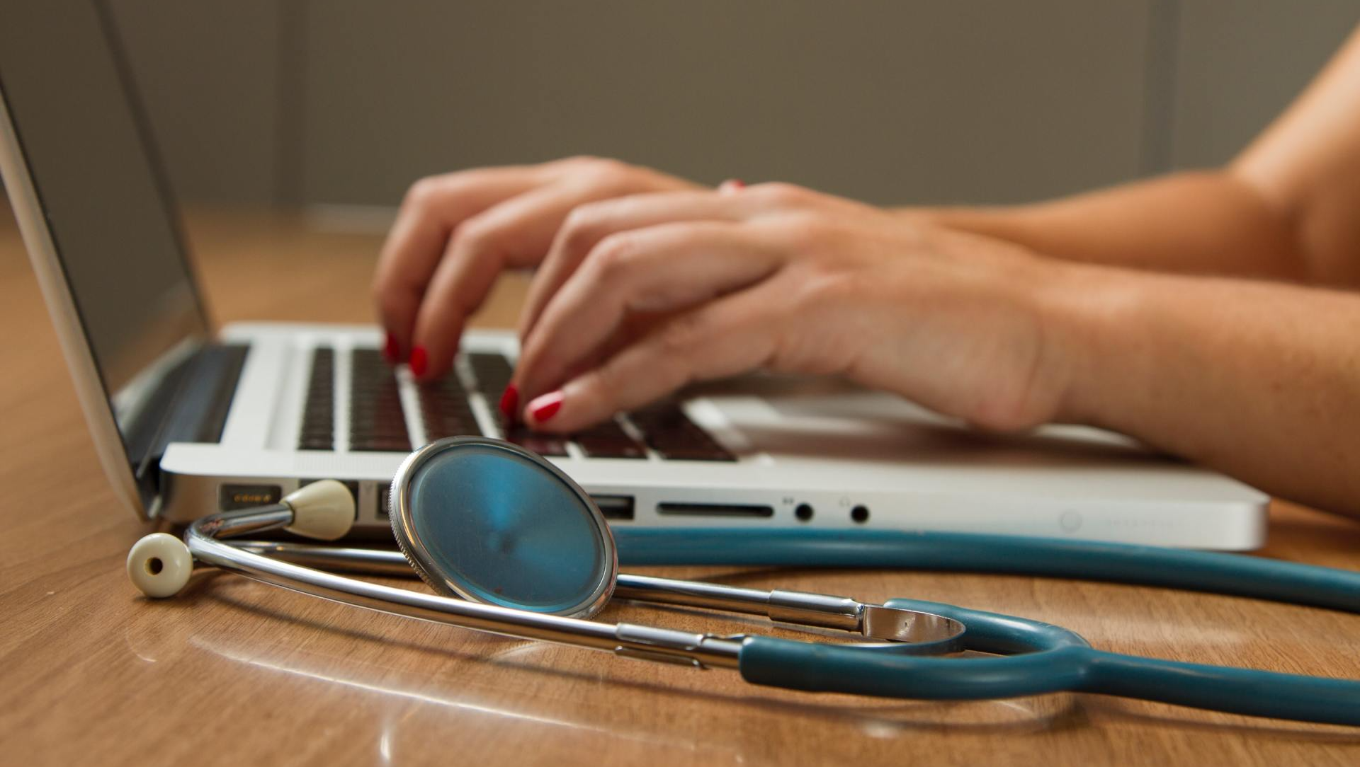A woman is typing on a laptop next to a stethoscope.