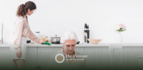 A senior sitting on a couch reading a bout while a caregiver cleans up the kitchen behind her