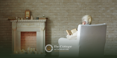 A senior sitting on a couch reading a book in front of a fireplace 