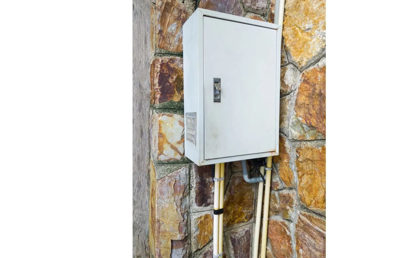 White electrical panel mounted on a stone wall with conduit and cables below.