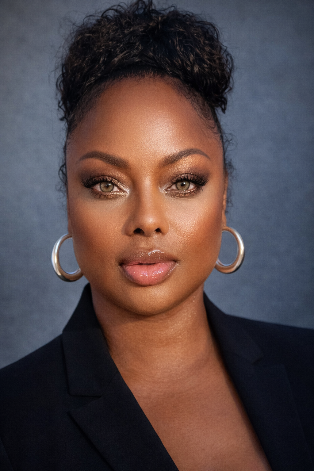 Headshot of a person with curly hair in a high bun, wearing gold hoop earrings and a black blazer against a grey backdrop.
