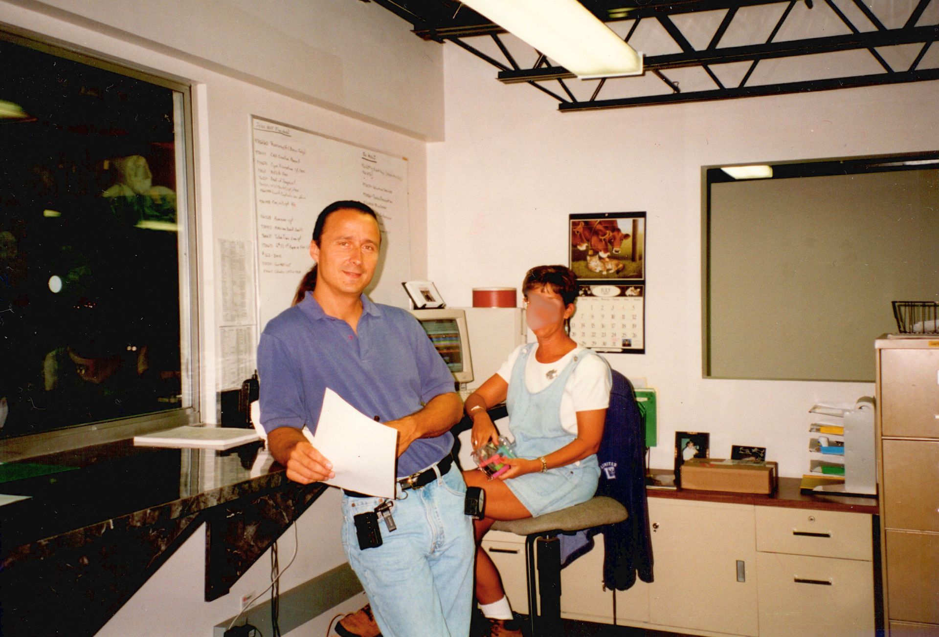 Two people in an office, one standing with papers and one seated at a desk, with shelves and a window in the background
