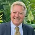 Smiling man in a navy suit and yellow tie, standing before lush green tropical leaves.