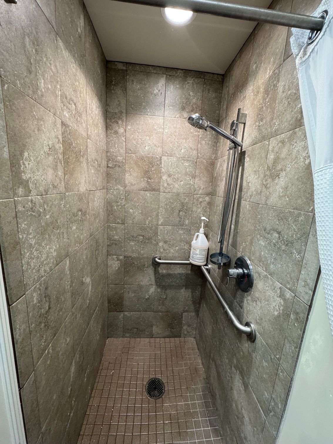 Small tiled shower stall with wall-mounted showerhead, grab bar, and soap shelf.