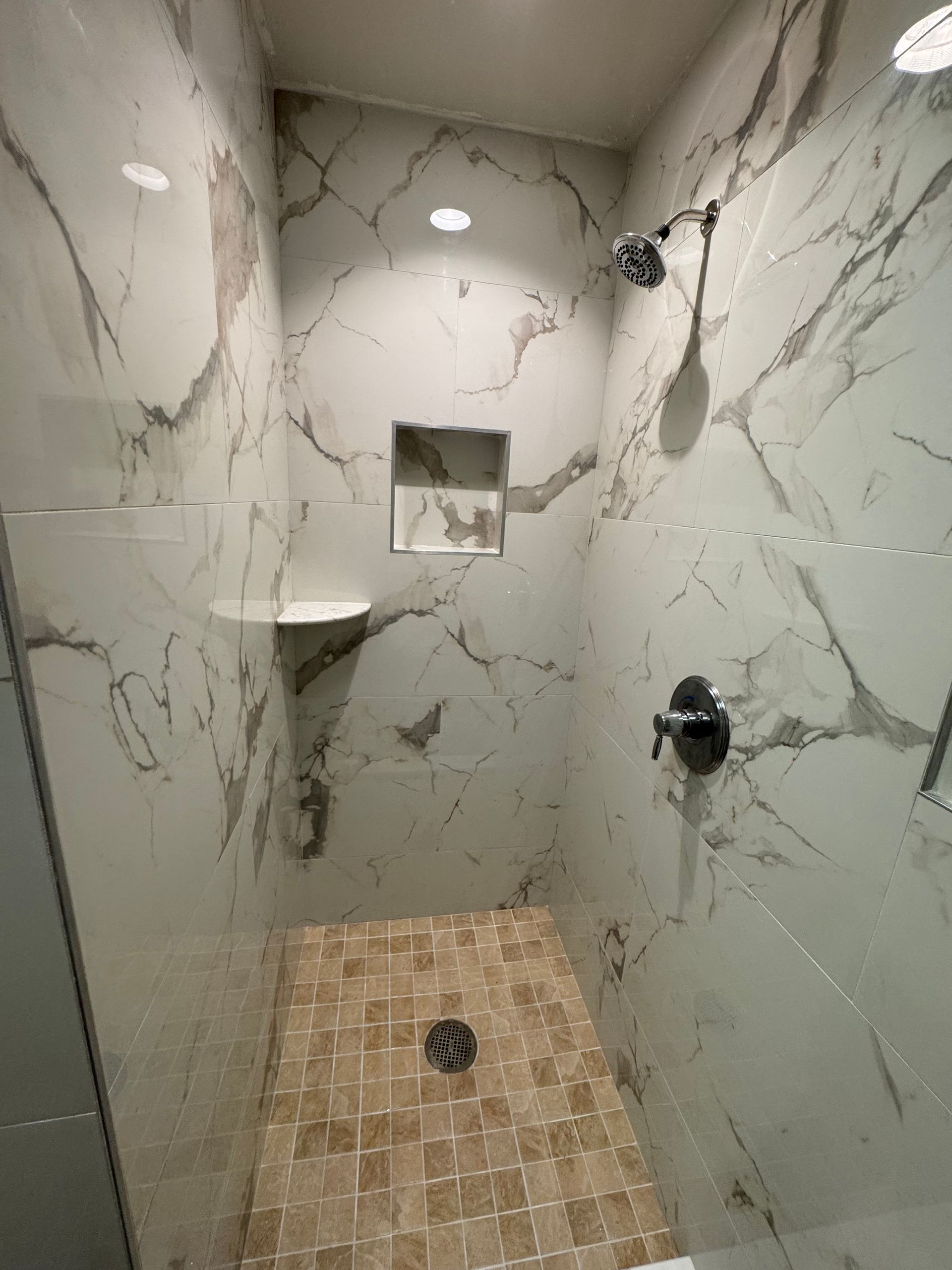 Narrow tiled shower stall with marble-pattern walls, built-in shelves, and drain in the floor