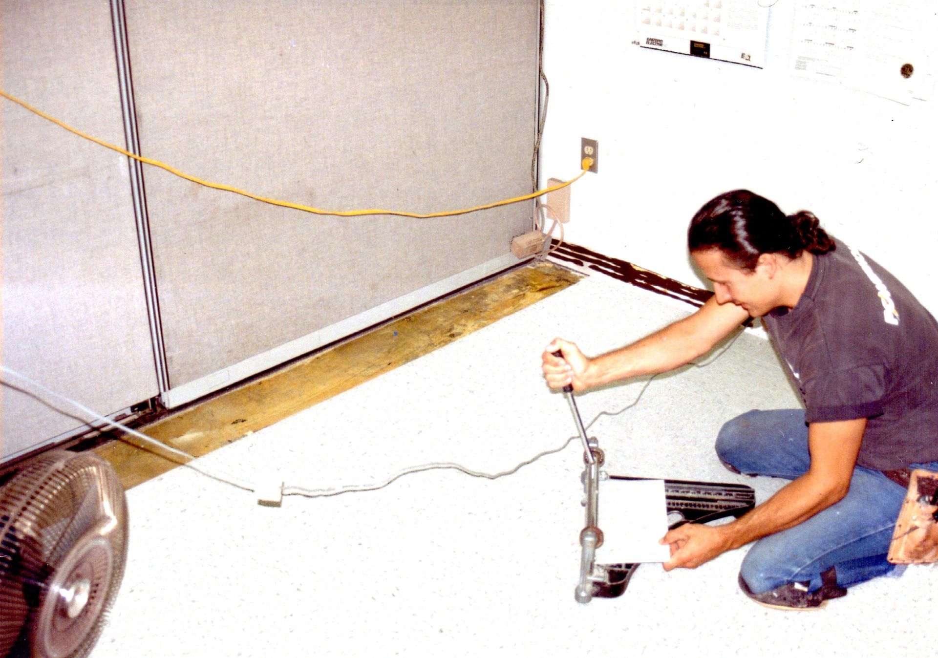 Man kneeling in a garage, repairing a car bumper near a floor fan and coiled cables.