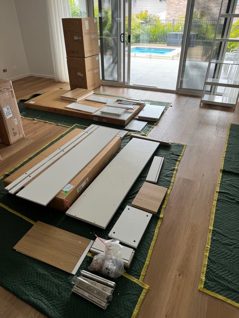 Stacks of flat-pack boxes and boards on a floor in a bright room near a sliding glass door.