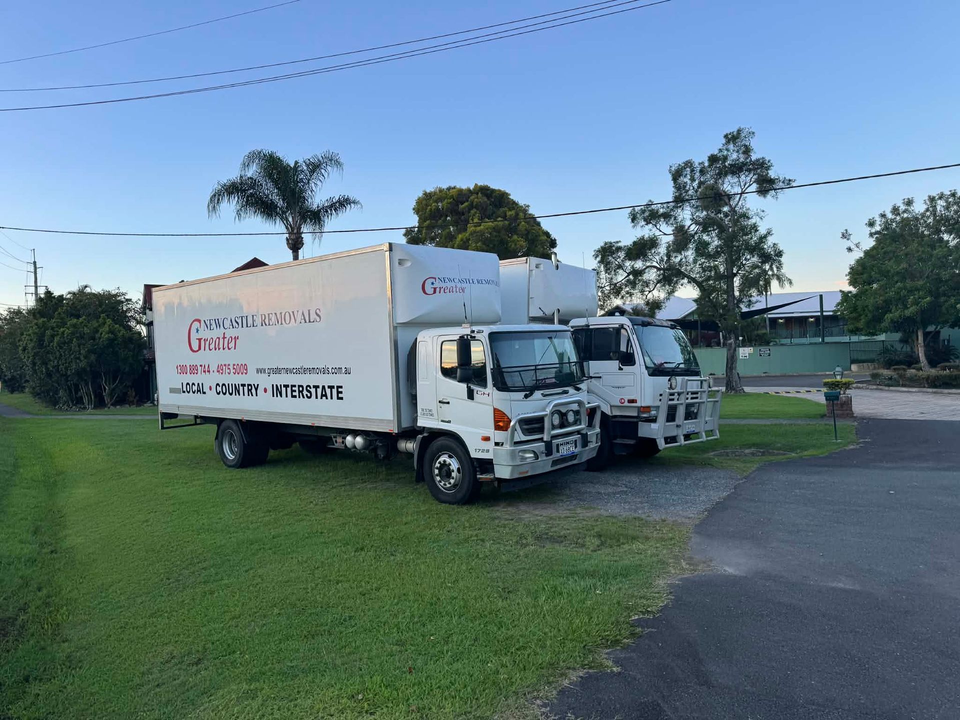 Two white moving trucks parked on a grassy roadside under a blue sky