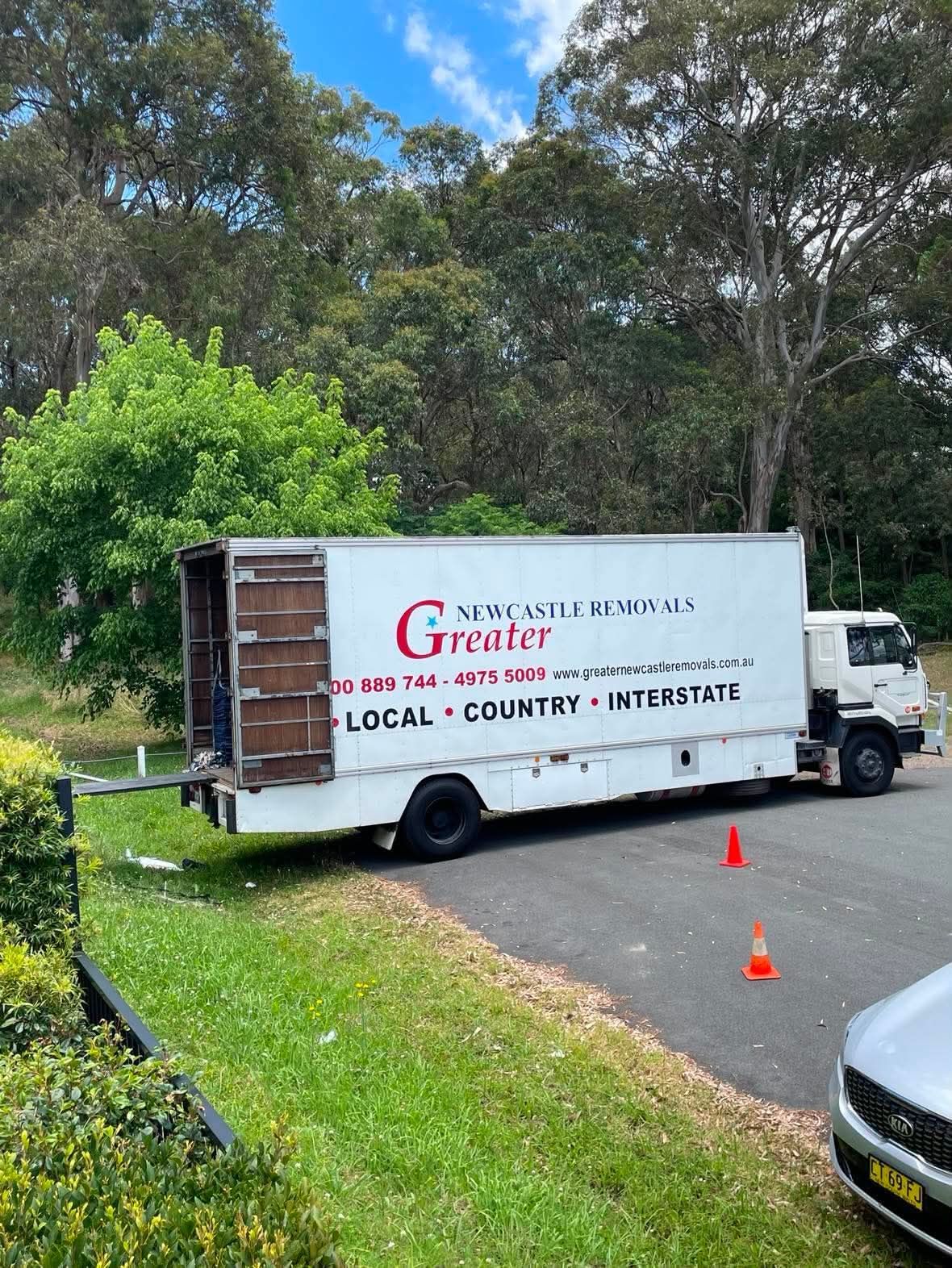 White moving truck with logo parked on a driveway, with orange cones and trees in the background