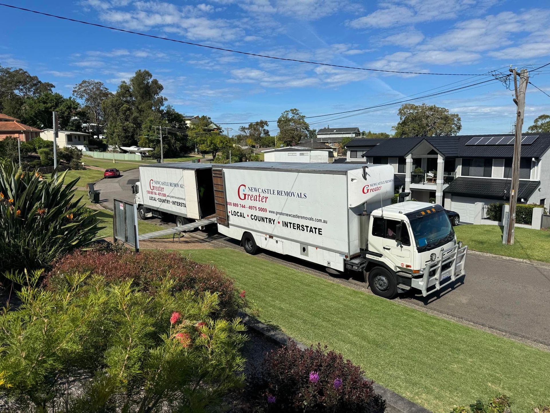 White moving truck on a suburban street beside a house and green lawn under a clear blue sky.