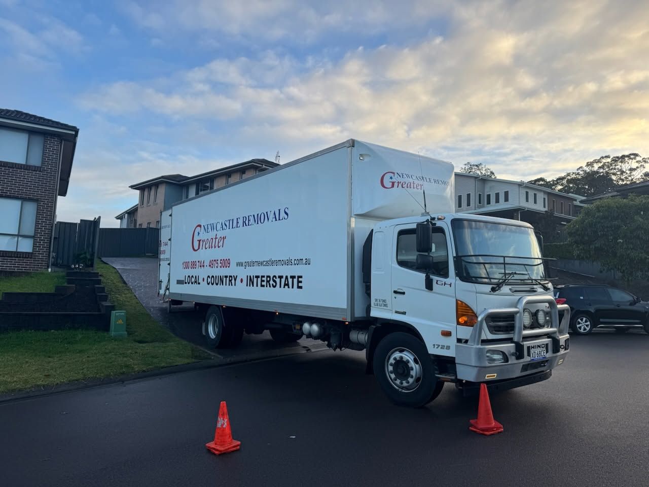 White box truck on a street with orange cones, parked outside buildings under a cloudy sky.