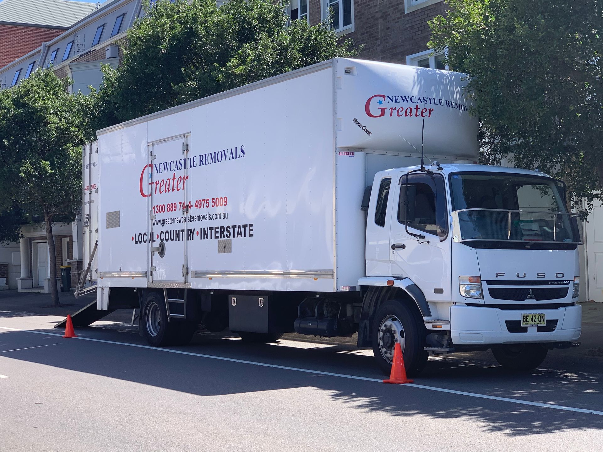 White moving truck parked curbside with orange cones on a sunny street.