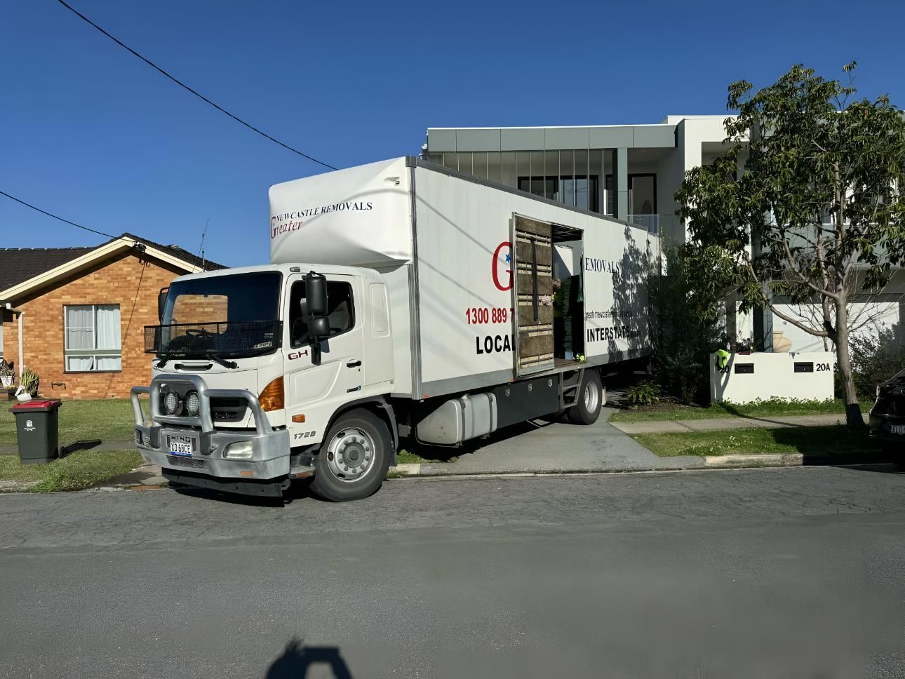 White moving truck parked on a residential street beside houses and trees under a clear blue sky