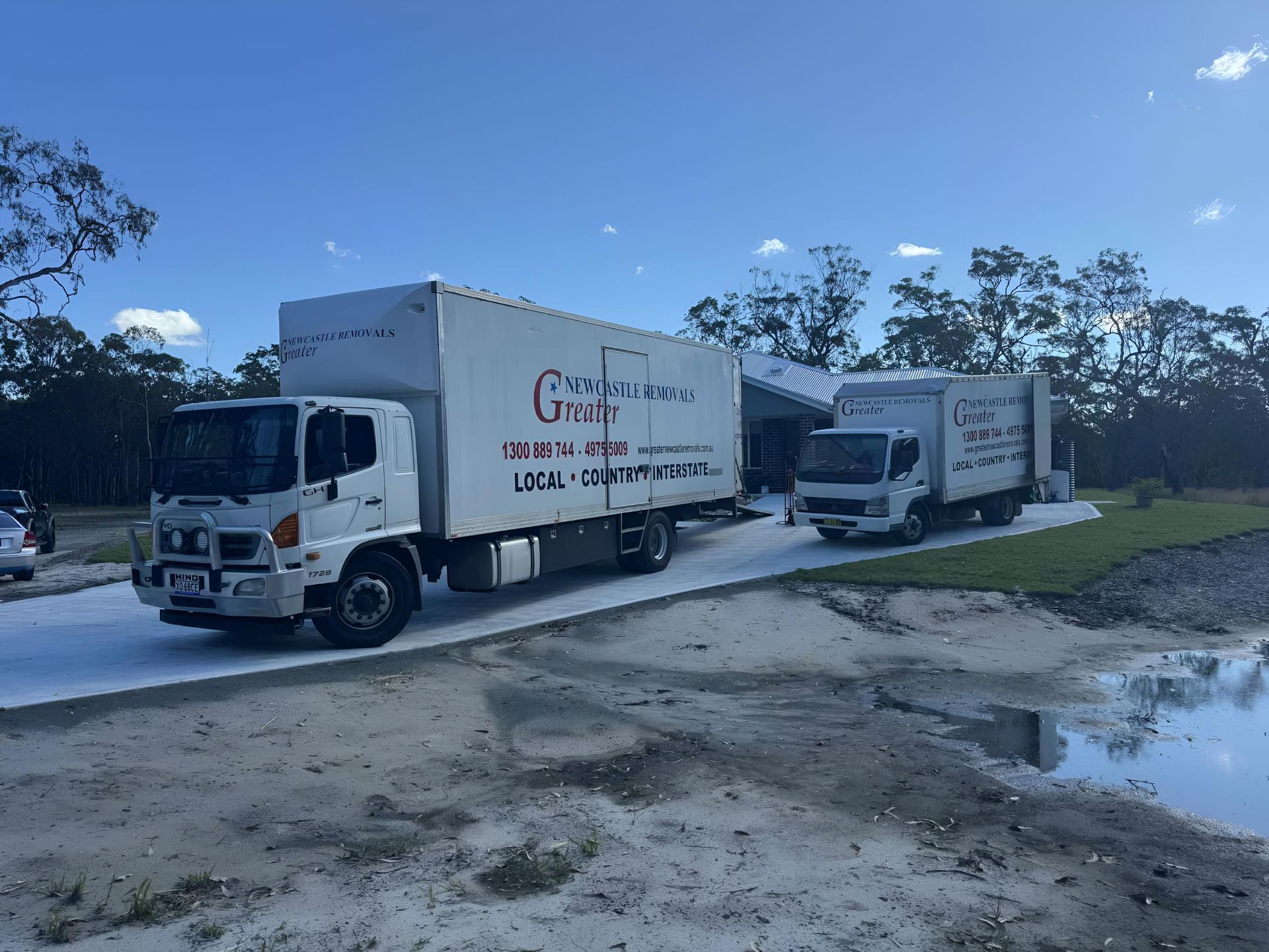 Two white box trucks parked on a dirt road beside a puddle under a blue sky