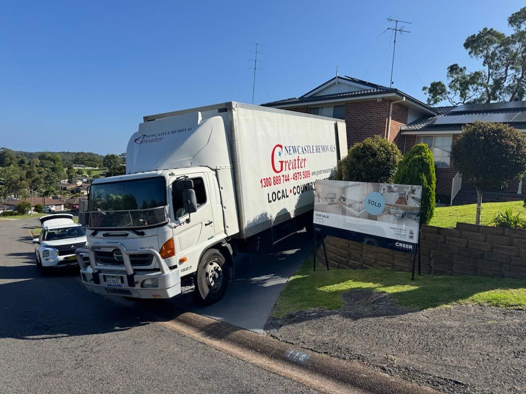 White moving truck parked by a suburban house with a for-sale sign on the lawn.