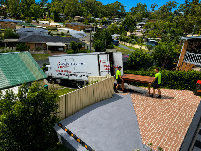 Two workers unload a truck on a brick driveway beside a house and hedge.