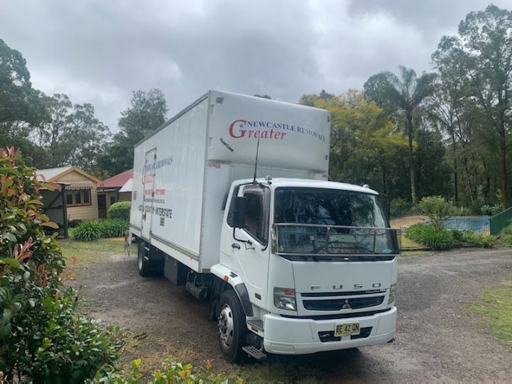 White delivery truck parked on a gravel road beside a house, with trees and cloudy sky in the background.