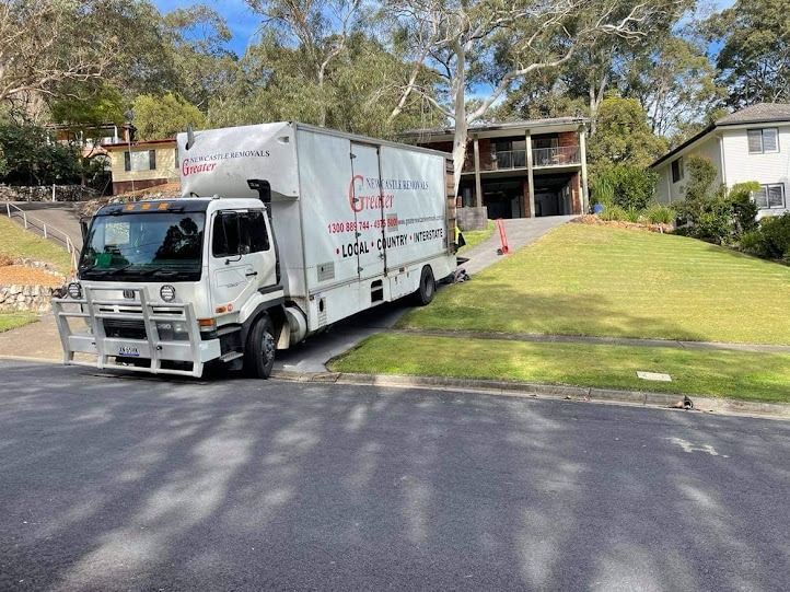 White moving truck parked on a grassy slope outside a house with a brick apartment building behind it
