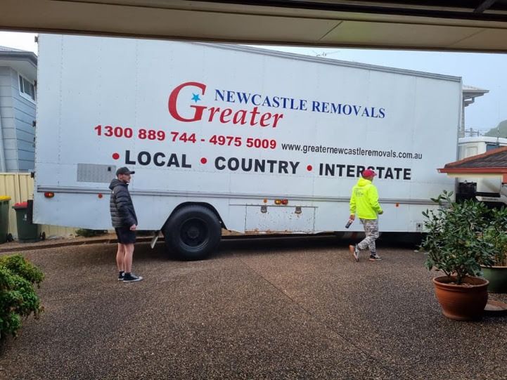 Two people standing beside a Newcastle Removals truck in a driveway, with potted plants nearby.