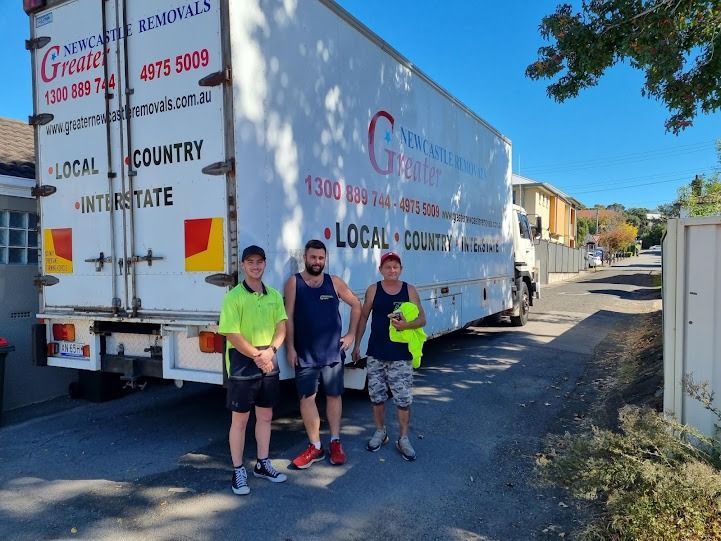 Three people standing by a large moving truck on a sunny street
