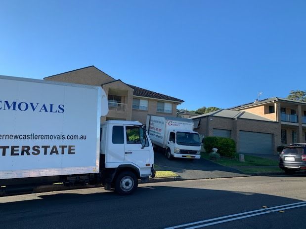Moving trucks parked outside suburban houses on a sunny residential street.