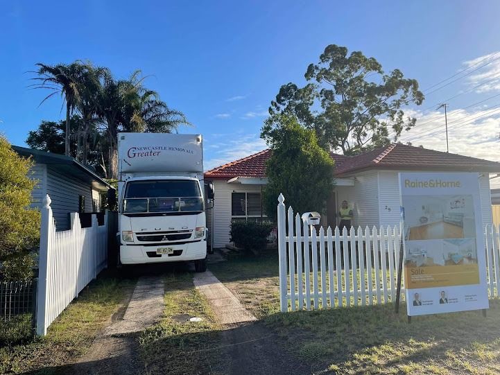 White moving truck parked in a driveway beside a suburban house with a white picket fence.