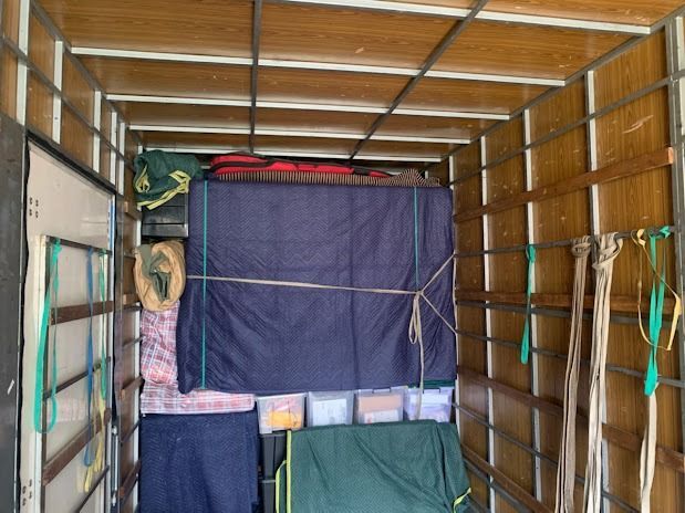 Inside a truck cargo area, a large blue tarp-covered item is strapped in place with storage bins below.