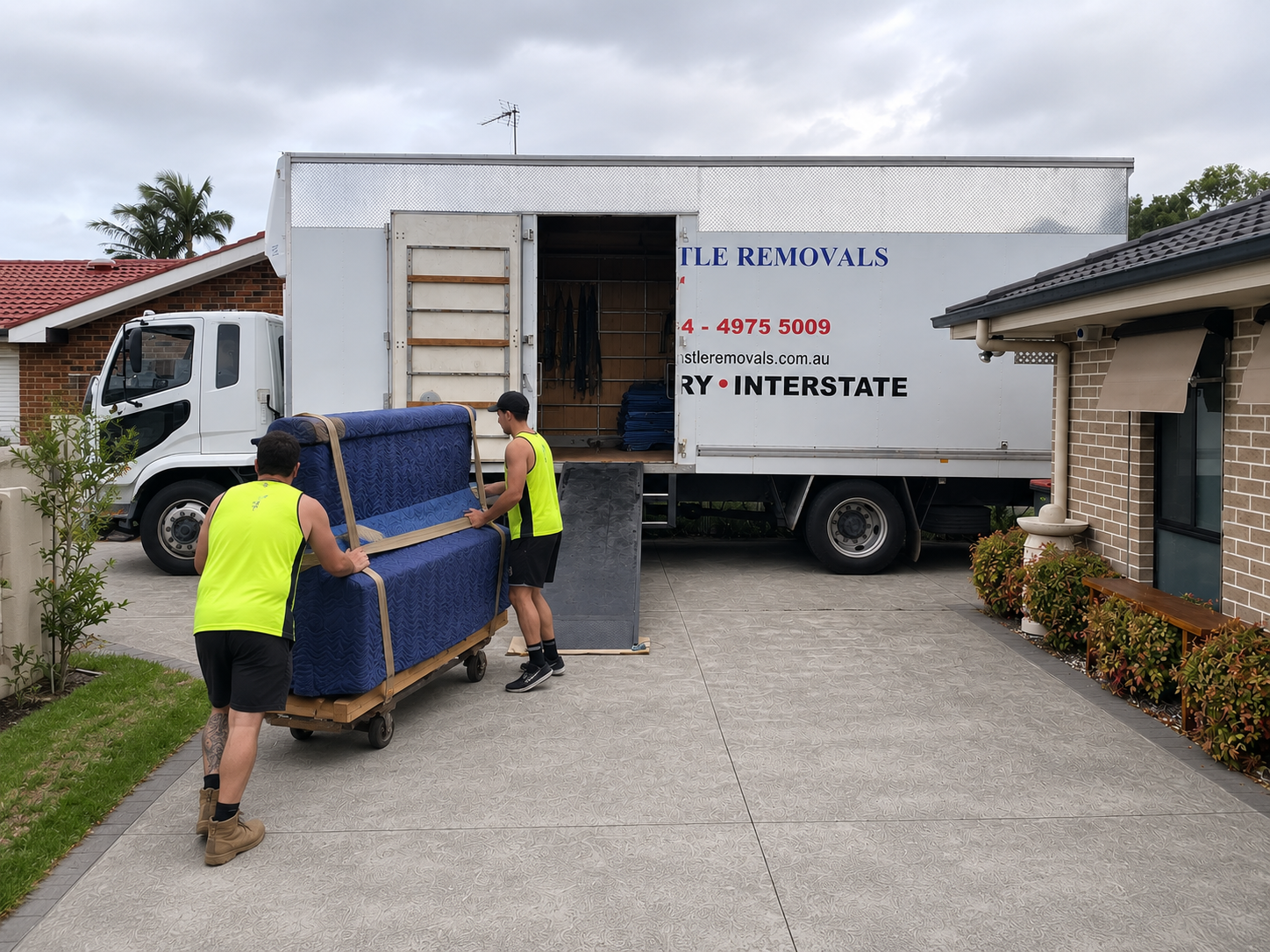 Two workers in neon vests unload a blue sofa from a moving truck in a driveway.