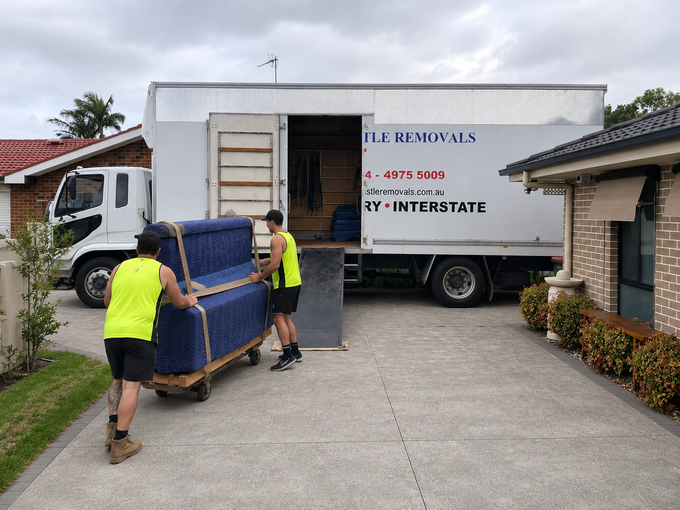 Two movers loading a blue couch into a white moving truck in a residential driveway.