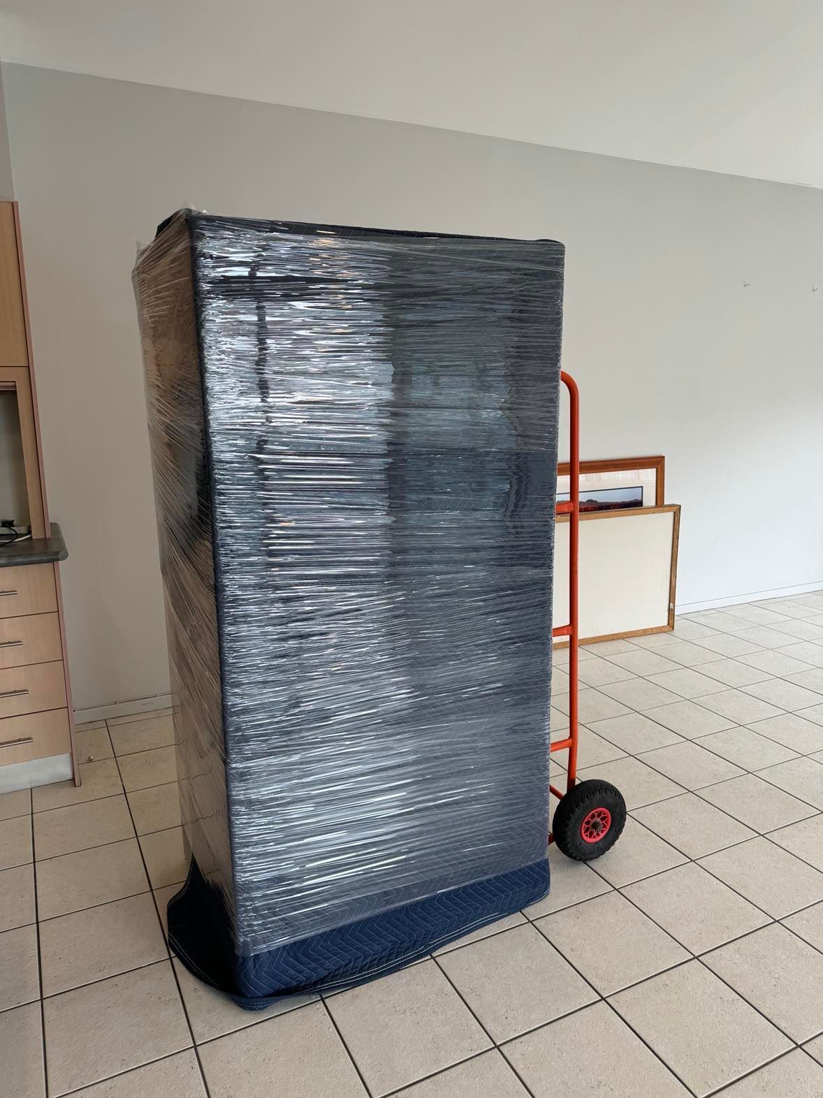 Tall black-wrapped pallet on a red hand truck in a tiled room
