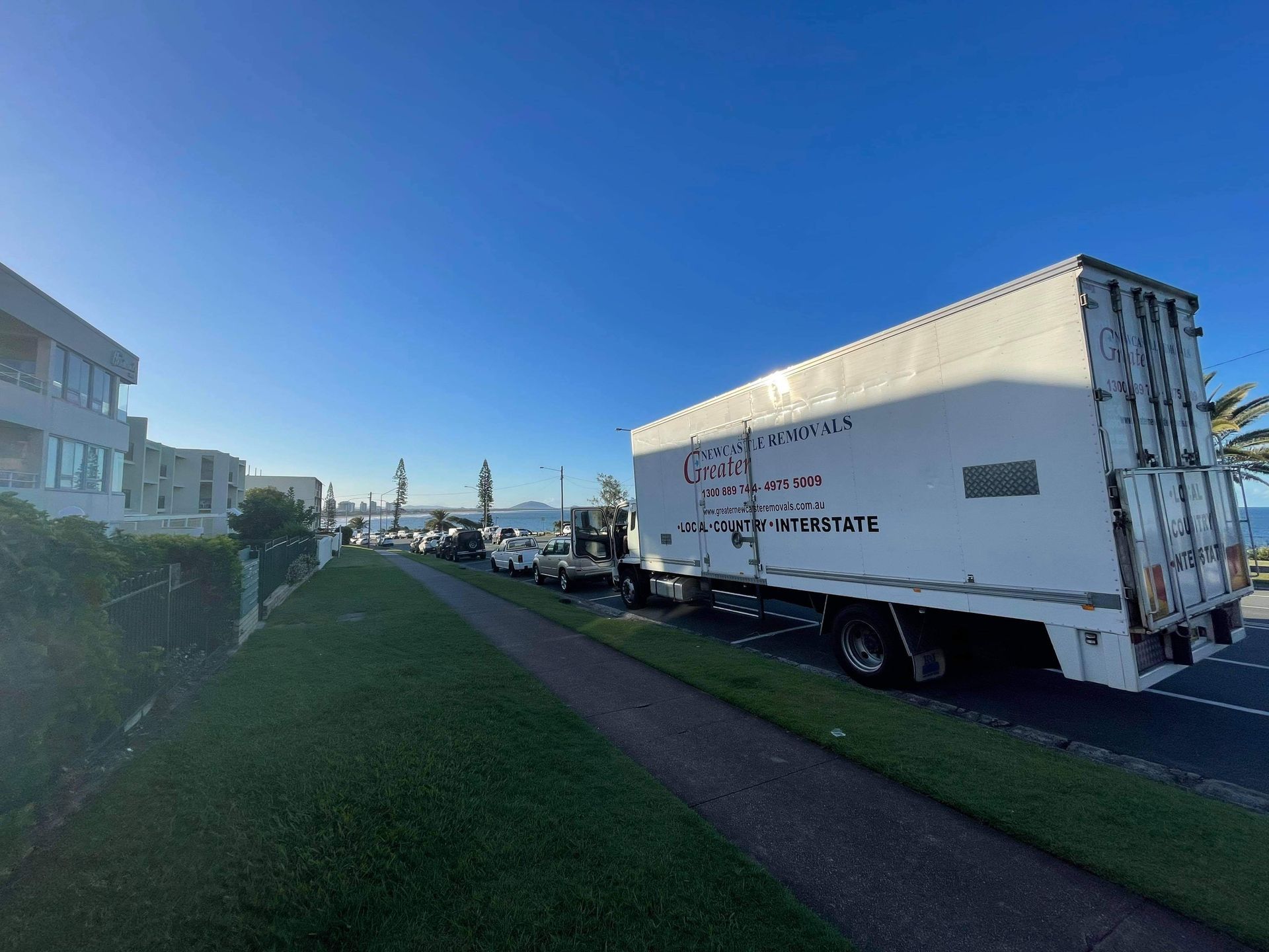 Large white box truck parked along a grassy path beside buildings under a clear blue sky