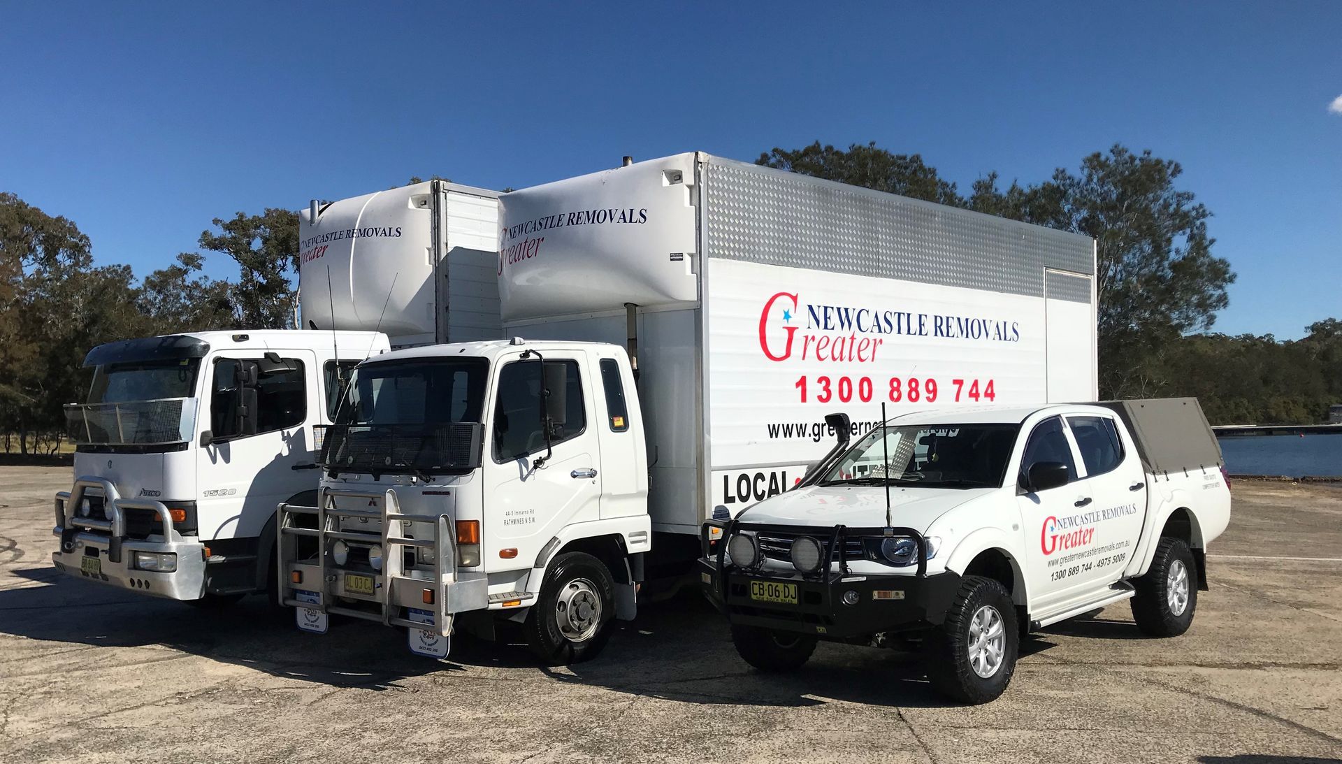 Three white work trucks parked on gravel, one with company signage, under a clear blue sky