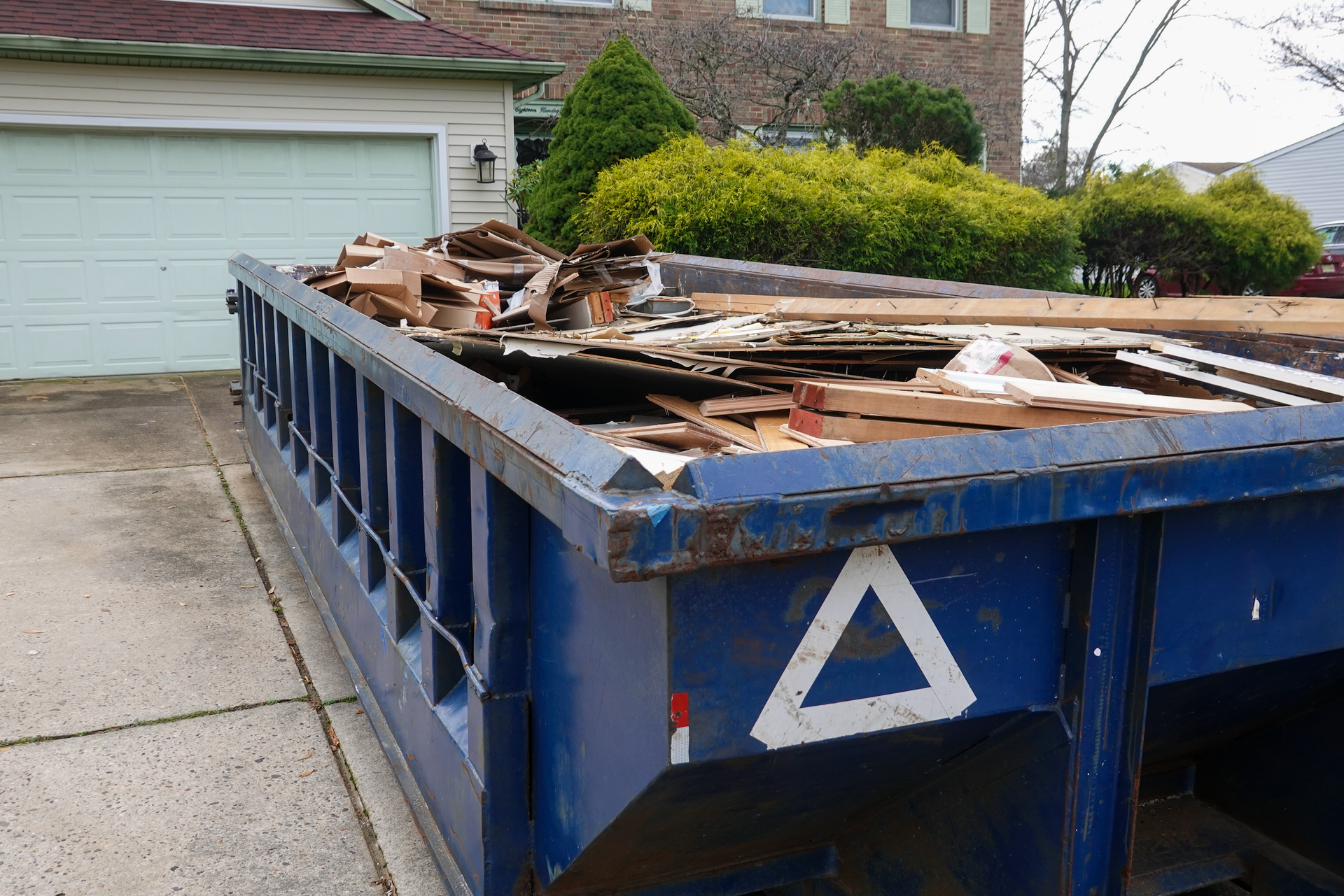Blue dumpster overflowing with construction debris, parked on a driveway in front of a house.