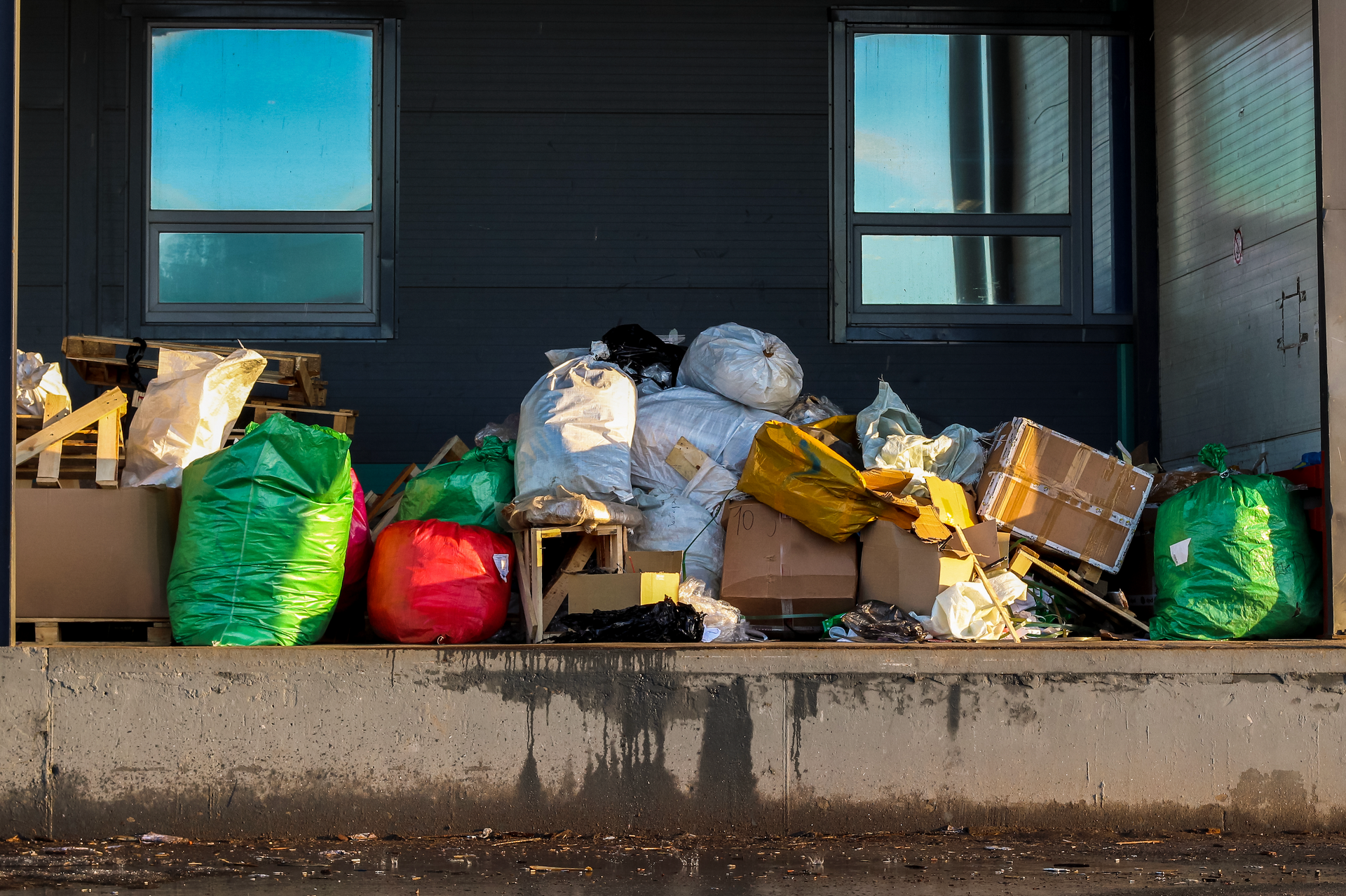 Trash bags and boxes piled on a loading dock against a dark building, two windows above.