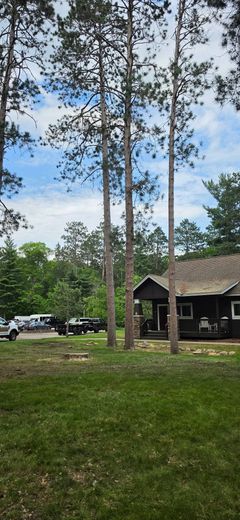 Tall trees in a grassy yard beside a dark cabin under a partly cloudy sky.