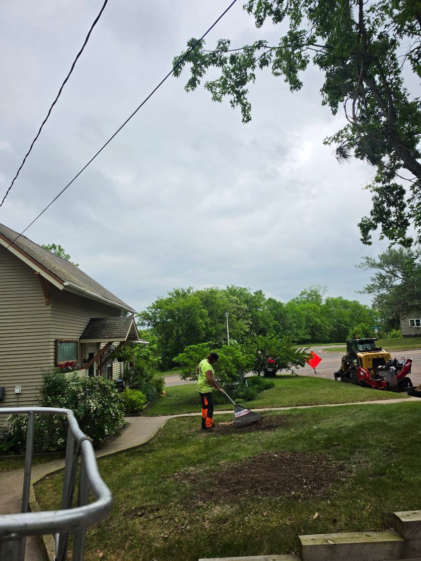 Workers using a string trimmer in a grassy yard beside a house and parked vehicles under cloudy skies