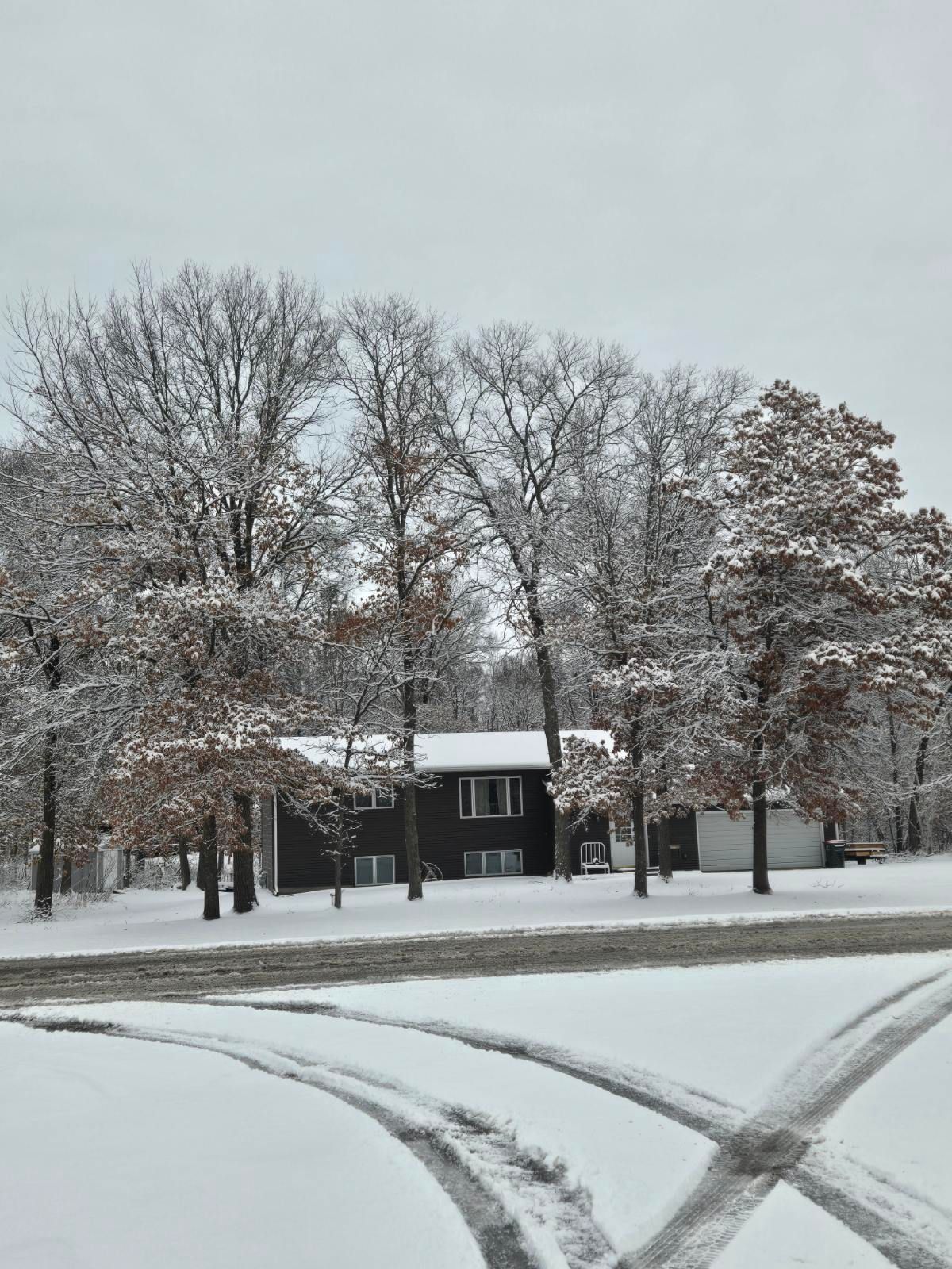 Snowy street with tire tracks in front of a house and bare trees