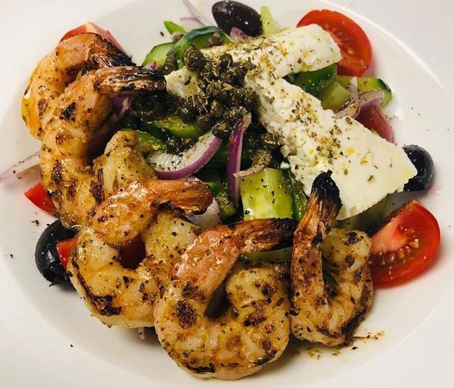 Grilled shrimp salad with feta, olives, tomatoes, cucumber, and red onion on a white plate