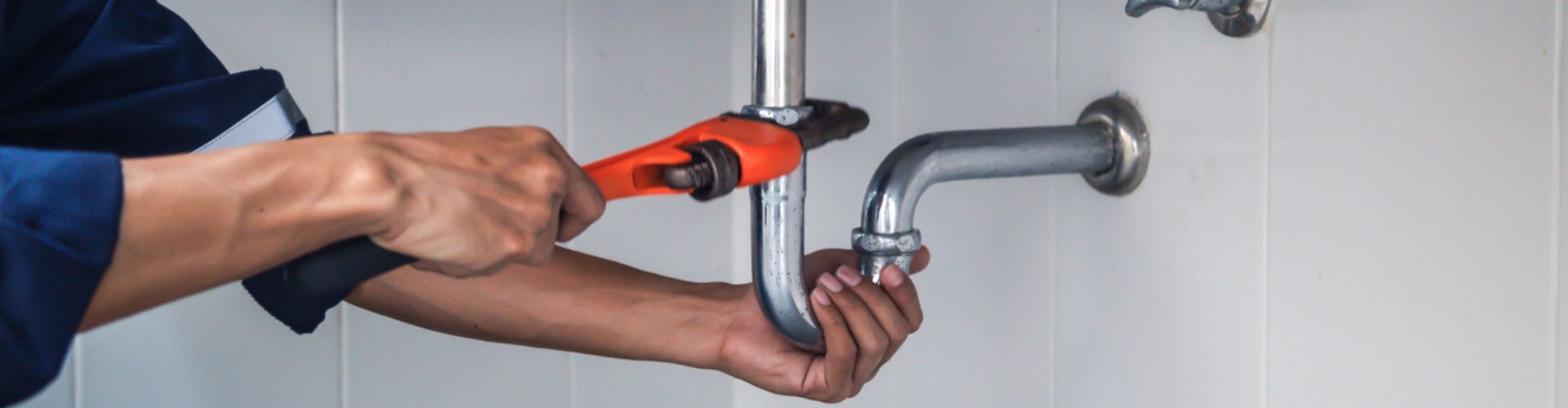 Plumber using a wrench to work on a pipe under a sink, with white tile background.