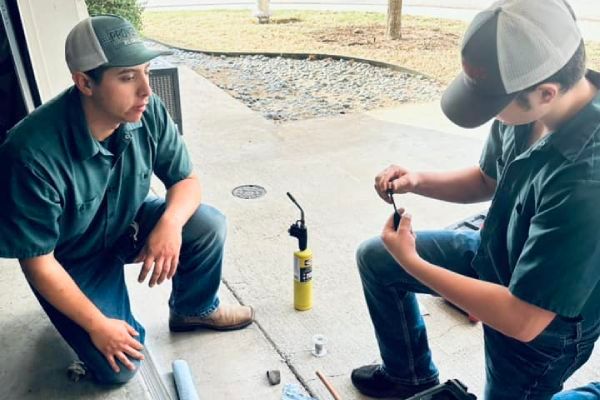 Two workers kneeling, soldering a small pipe, with a propane torch, on a concrete surface.
