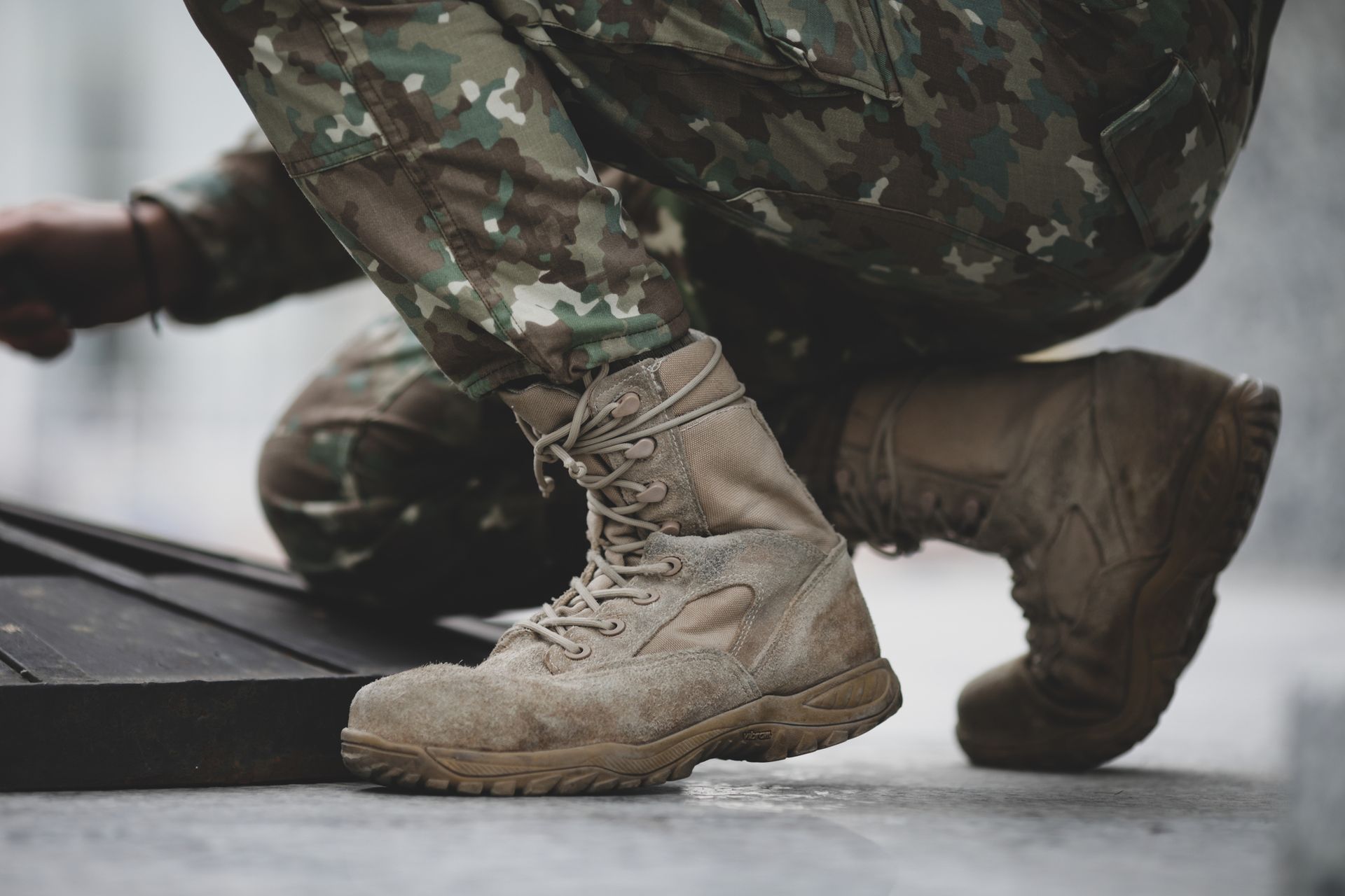 Camouflage-clad person crouching in tan combat boots on a wet surface