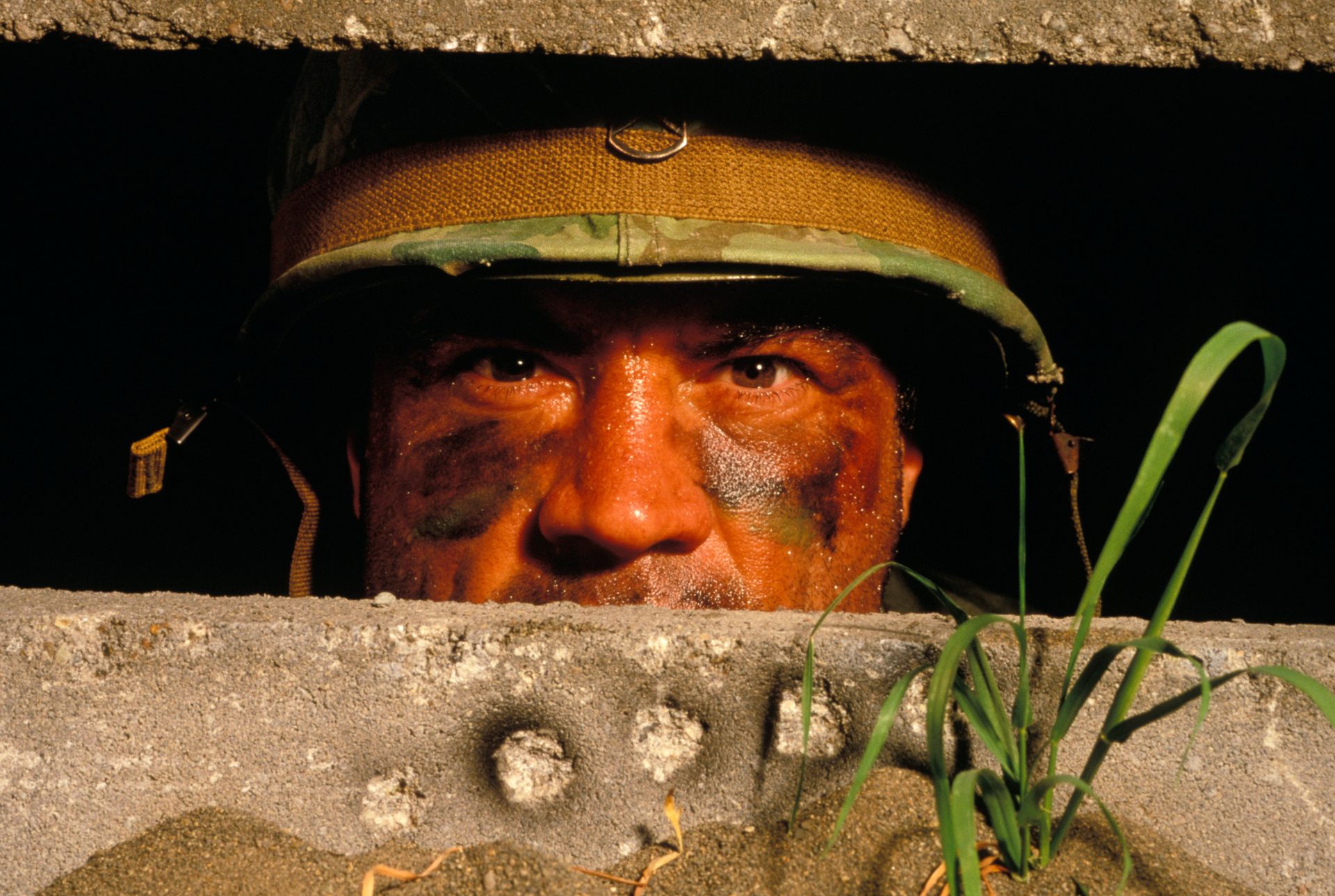 Soldier peering over a concrete barrier, wearing a helmet in warm sunlight