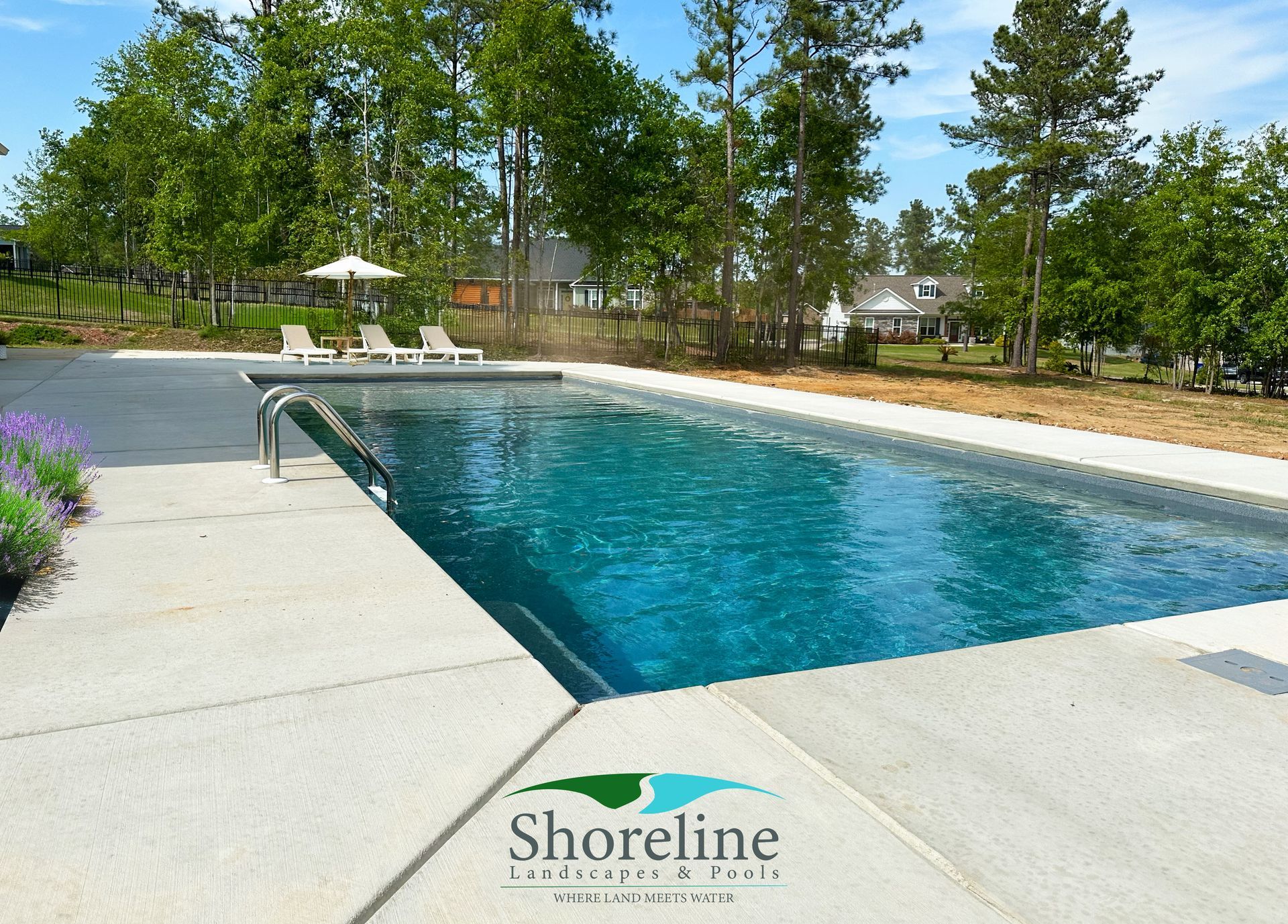 Rectangular in-ground pool surrounded by concrete, with lounge chairs and a parasol in the background. Shoreline Pools logo visible.