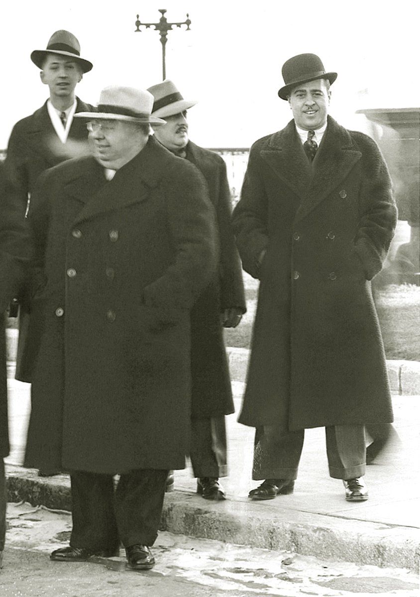 A group of men wearing hats and coats are standing on a sidewalk